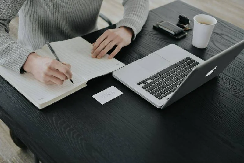 Person writing audit notes beside a laptop